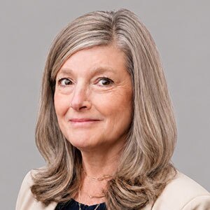 Middle aged woman with long blonde gray hair wearing a black top and beige blazer.