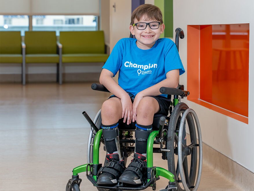 A smiling boy with glasses sits in a manual wheelchair, wearing a blue “Champion” t-shirt and leg braces.