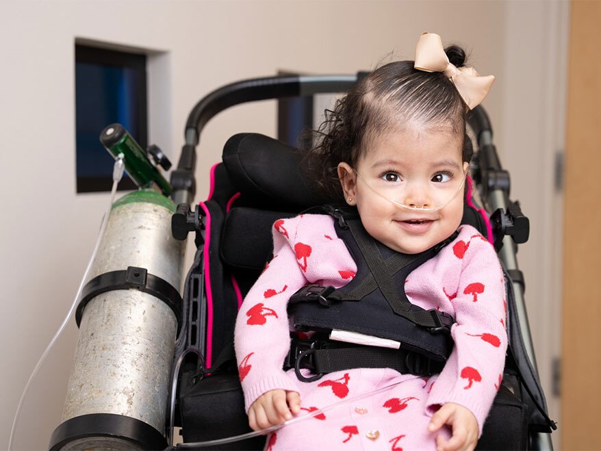 A young child with a nasal cannula sits in a medical stroller attached to an oxygen tank.