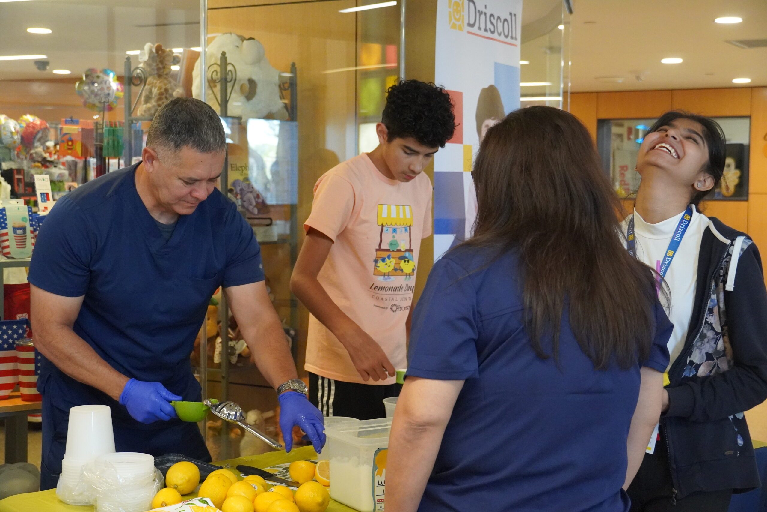 Luke and a man in dark blue scrubs make lemonade for a woman at a booth in a mall.