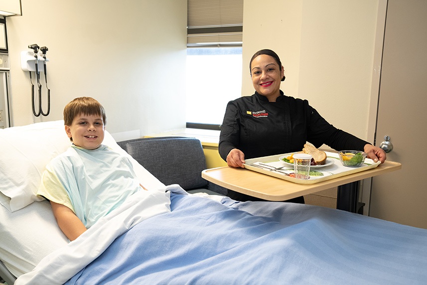 Boy in a hospital bed while nurse is serving him a lunch tray.