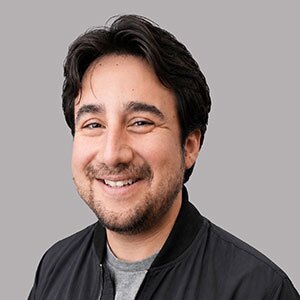Smiling man with short black hair wearing a gray t-shirt under a black button up collard shirt.